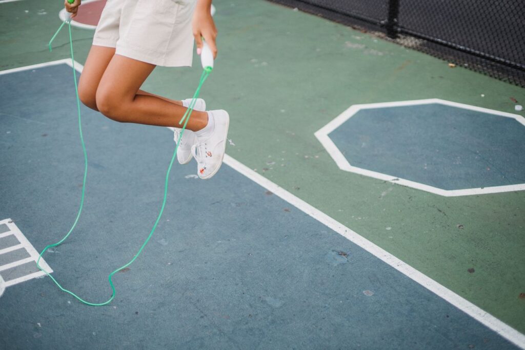 A girl playfully jumping rope on a painted outdoor playground surface, wearing casual shorts and sneakers.
